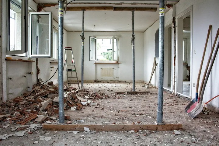 Gutted room with walls and floors torn out, debris piled on the subfloor, and temporary steel shoring posts holding up the ceiling